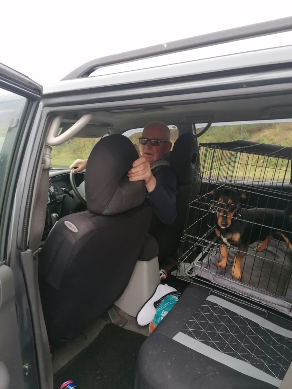 Older man with short, light gray hair and glasses, seated behind the driver's seat of a dark gray SUV. He's wearing a dark-coloured, long-sleeved shirt and appears to be adjusting or holding the headrest of the passenger seat. His expression is somewhat serious, even slightly strained. In the back of the SUV is a dark-colored dog, possibly a mix breed, contained within a wire dog crate. The dog is positioned towards the back of the crate, facing slightly towards the camera. It seems relatively calm. The SUV's interior is largely dark-coloured, with black seat covers. There are various items scattered in the back, including a pair of white sneakers and what appears to be a small, brightly colored item (possibly a bag or towel). A partially visible, crushed aluminium can sits on the car floor near the driver’s side.