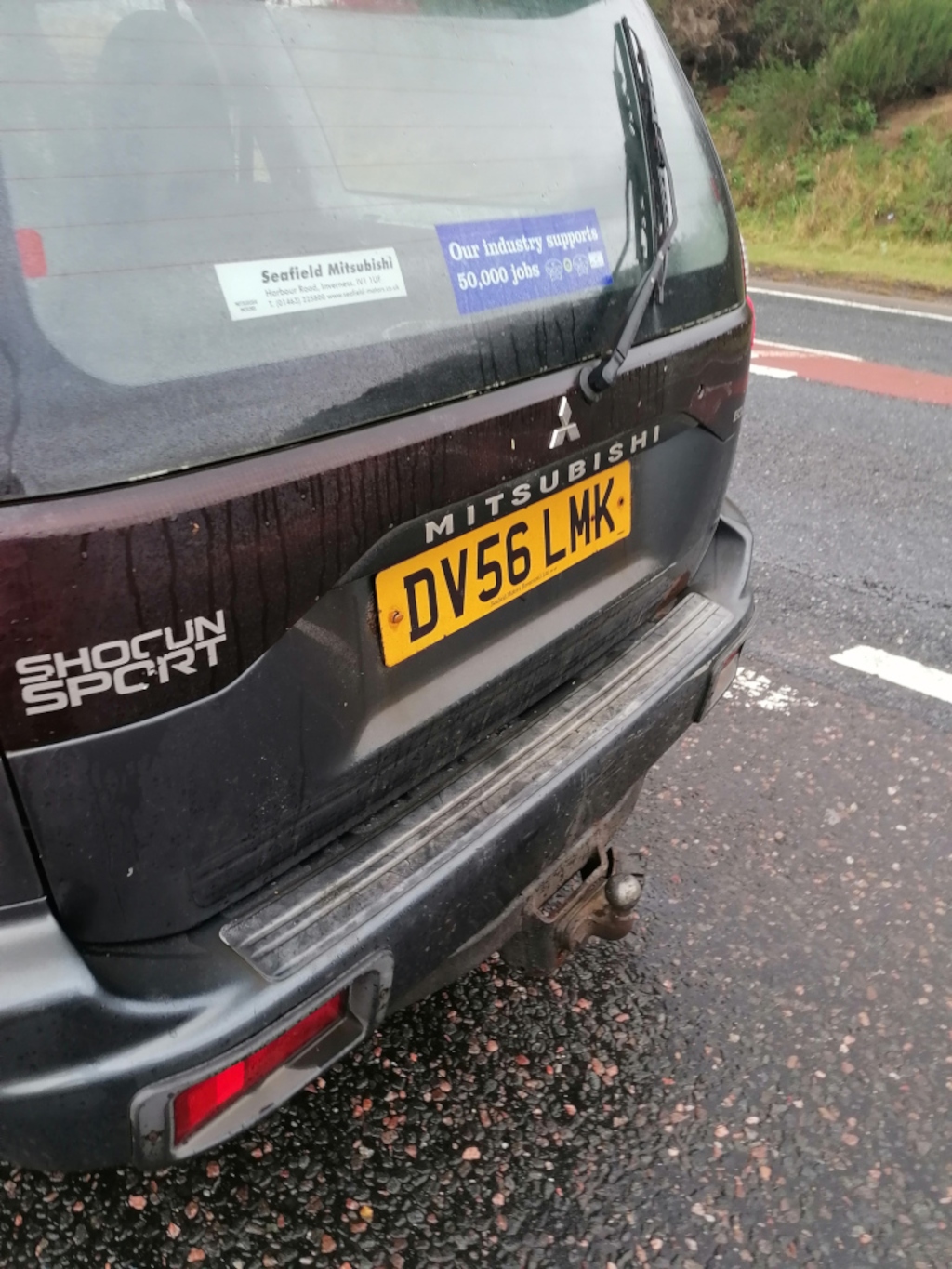 Rear of a dark-grey or brownish-grey Mitsubishi Shogun Sport SUV. The vehicle is parked, slightly angled to the right, on a tarmac road. The license plate, DV56 LMK, is prominently displayed in a bright yellow rectangle. Several stickers adorn the rear window: one advertising Seafield Mitsubishi, a dealership in Inverness, and another highlighting the industry's contribution of 50,000 jobs. A tow hitch is visible beneath the rear bumper. The car appears to be slightly wet or dusty. The setting is outdoors, on a road with a faint white line marking a possible lane or pedestrian area. In the background, there’s blurred greenery suggesting trees and possibly a woodland area. The road is dark grey asphalt, with small stones and gravel visible closer to the camera. A section of light red/pink tarmac can be seen further away.