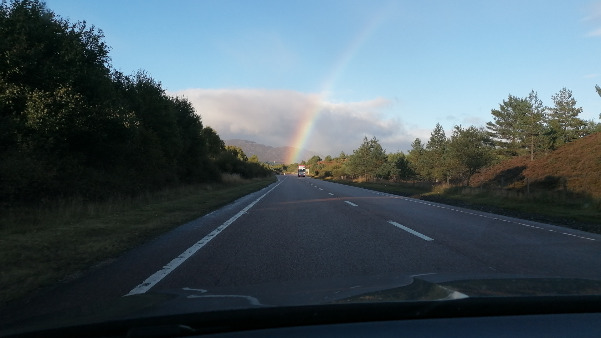 A scenic view from inside a car driving down a highway. A vibrant rainbow arches across the sky in the distance, above a landscape of dark green trees and rolling hills. The road ahead is fairly straight, and the sky is a clear, light blue. The overall impression is one of tranquil beauty and open space.