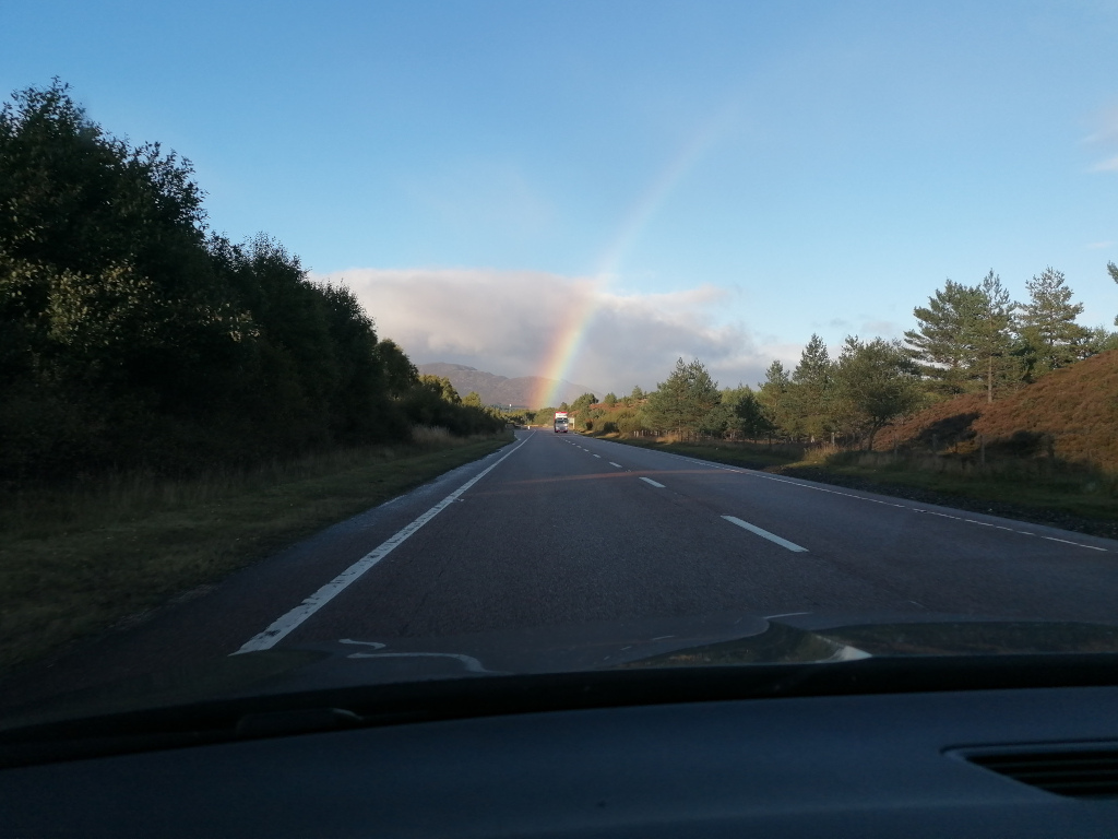Scenic view from inside a car driving down a highway. A vibrant rainbow arches across the sky in the distance, above a landscape of dark green trees and rolling hills. The road ahead is fairly straight, and the sky is a clear, light blue. The overall impression is one of tranquil beauty and open space.