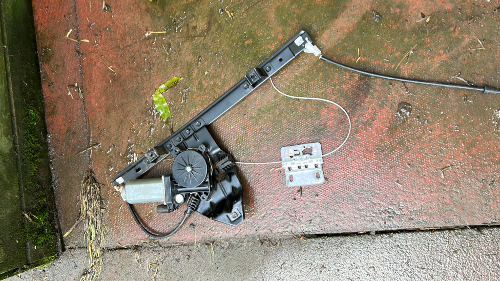Disassembled car window regulator mechanism lying on a wet, reddish-brown ground. The parts include the regulator motor, the track, and associated cables and brackets. The background suggests an outdoor setting, possibly a repair or disposal area.