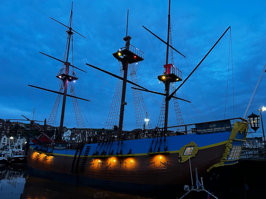 Large, wooden pirate ship at dusk or night. It's elaborately decorated, with numerous lights illuminating the vessel against the twilight sky. The ship appears to be docked at a harbor, with other boats and buildings visible in the background. A banner on the ship indicates that food and drink are served all day. The overall feeling is one of adventure and possibly a tourist attraction.
