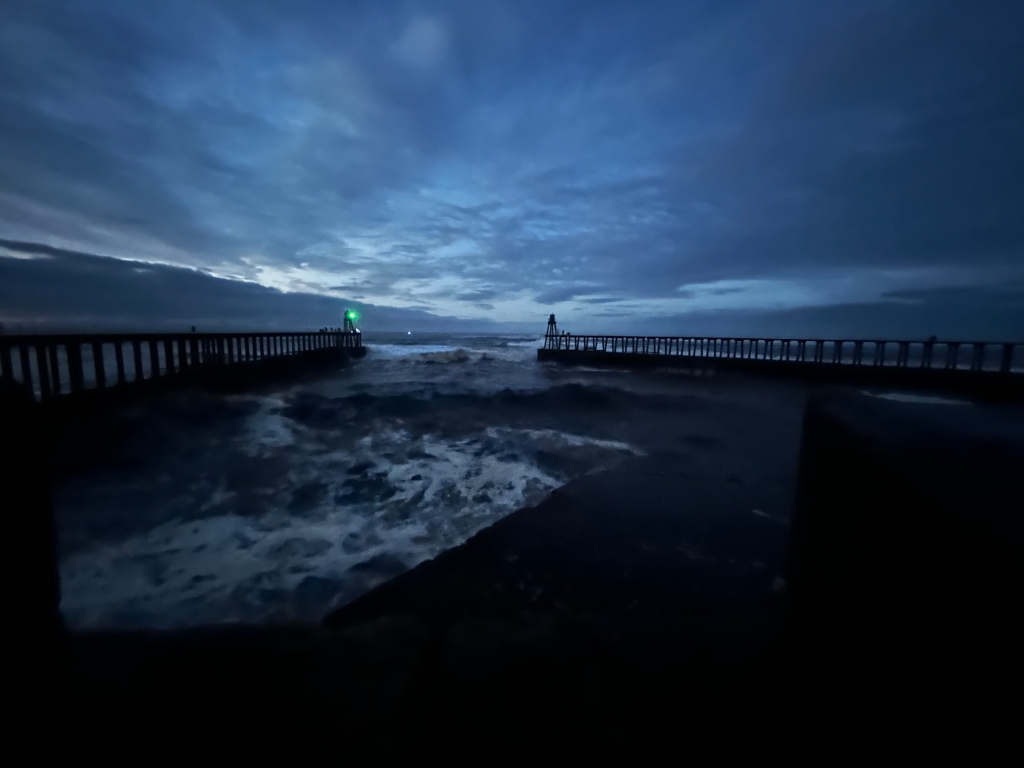 Dark, moody scene at what appears to be a pier extending into a turbulent sea at twilight or dawn. The sky is a deep blue, filled with clouds. The pier is silhouetted against the water and sky, with a small light visible at its end. The water is rough, with visible waves and foam. The overall atmosphere is one of solitude and dramatic natural power.