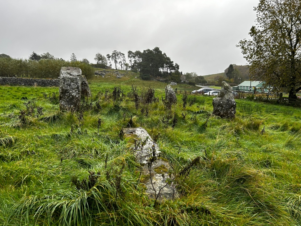 Grassy field with several large standing stones, possibly a stone circle or part of one. In the background, there's a low stone wall, some trees and a building, suggesting a rural or possibly archaeological site. The weather appears overcast.
