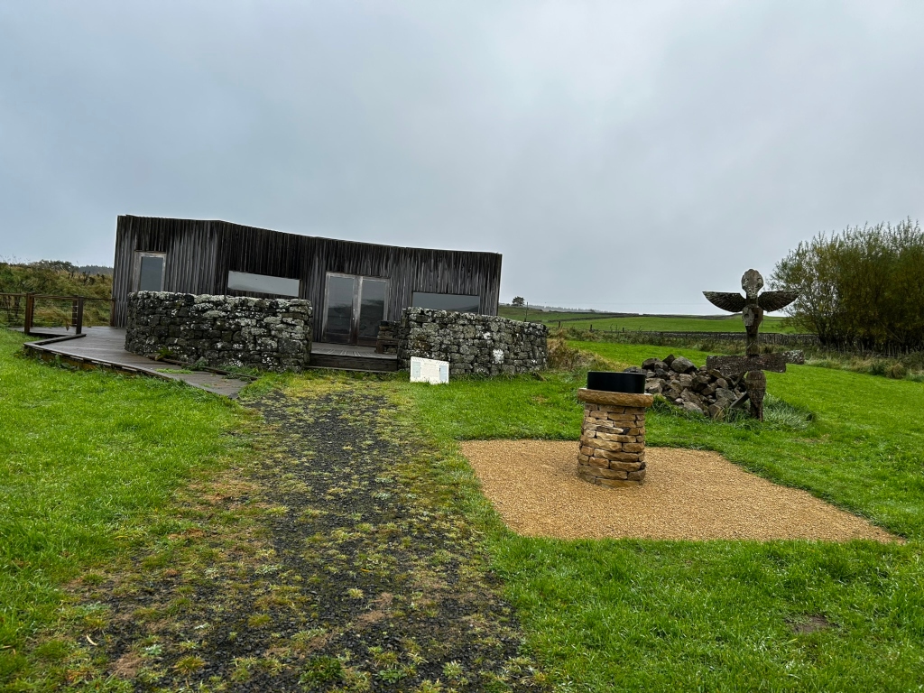 Modern, dark-wood building with a stone wall encircling its base. It's situated in a grassy field, and a short gravel path leads toward it. Near the building, there is a smaller stone structure resembling a pedestal, and further away stands a carved wooden sculpture that resembles a stylised bird or angel. The overall setting appears rural and possibly part of a nature reserve or similar location. The sky is overcast.