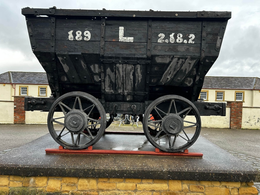 Weathered, dark-coloured wooden mining cart or wagon, prominently displaying the numbers 189 and 2.68.2 along with the letter L painted on its side. The cart rests on a low stone pedestal in front of a brick building, suggesting it's a historical artefact displayed outdoors. The overall impression is one of age and industrial history.