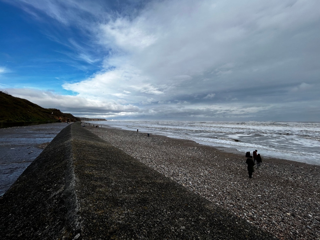 Long, dark grey concrete seawall stretching from the bottom left corner towards the middle distance, where it meets a pebbly beach. The beach extends to a choppy grey sea under a partly cloudy sky. A small group of people are visible on the beach near the seawall. The sky is a contrast of blue and grey, suggesting a somewhat stormy day but with patches of brighter sky. The overall impression is one of a bleak, yet dramatic coastal scene.