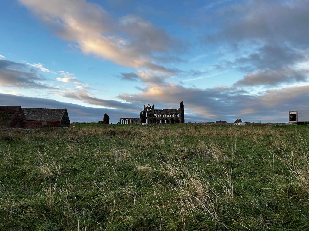 Landscape at dusk or dawn.  In the foreground is a field of tall grass. In the middle ground, there are a couple of smaller buildings to the left, and the ruins of Whitby Abbey are prominently featured in the center. A white vehicle is partially visible in the far right background. The sky is a mix of blue and clouds with some warm, pale hues near the horizon. The overall mood is serene and slightly melancholic, typical of a twilight scene featuring ancient ruins.