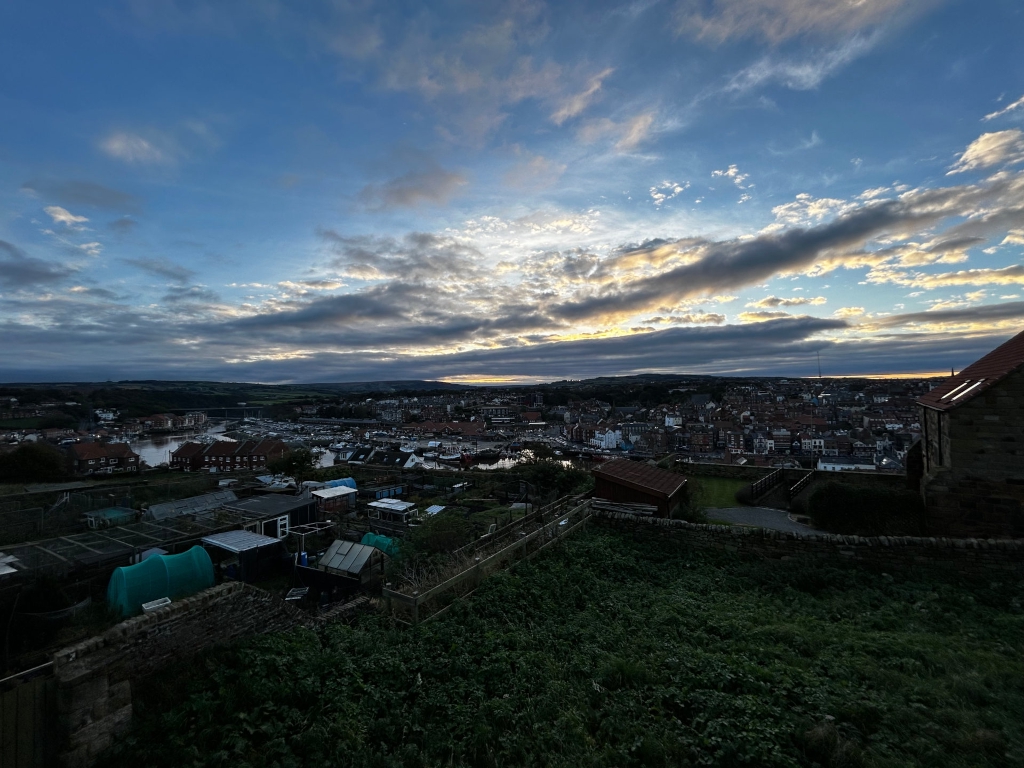 High-angle, long shot of a coastal town at sunset. In the foreground, there's a line of allotments or garden plots, nestled behind a low stone wall. Beyond this, the sloping land reveals a panoramic view of the town, its harbor densely packed with boats, and the surrounding landscape. The sky is a dramatic mix of dark clouds and streaks of lighter, yellowish clouds, typical of a sunset. The overall mood is serene yet slightly melancholic, given the darkening sky and the somewhat subdued lighting on the town below. There's a sense of quiet community in the juxtaposition of the personal, intimate scale of the allotments with the broader view of the inhabited town.