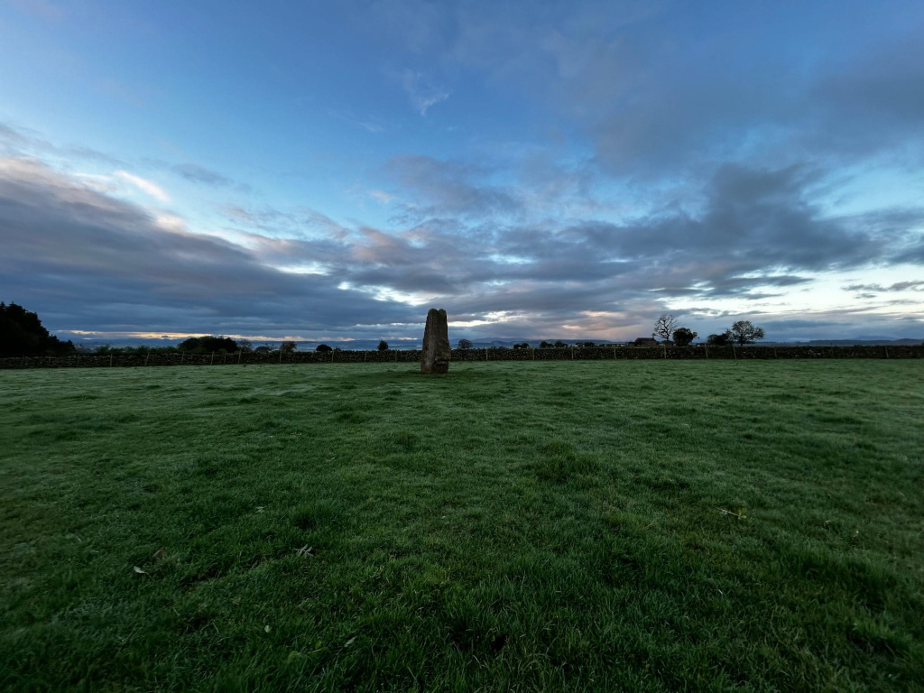Single standing stone in a grassy field, enclosed by a low stone wall. The sky is a blend of blue and grey clouds, suggesting either dawn or dusk. The overall mood is serene and somewhat mysterious.