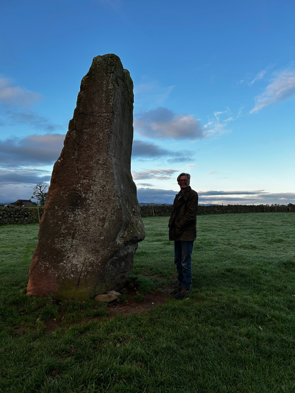 Charlie standing next to a large, upright stone, possibly a standing stone or menhir. The stone is tall and somewhat weathered, appearing ancient. He is dressed casually and seems to be observing or admiring the stone. The setting is a grassy field under a clear, mostly blue sky with a few clouds.