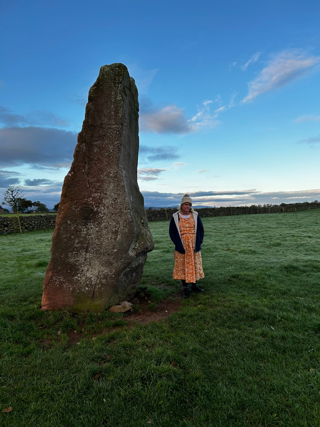 Leonie standing next to a large standing stone in a grassy field. The sky is clear and blue, with some clouds. She is wearing a long dress, a jacket and a hat. The standing stone appears ancient and weathered. The overall impression is one of serenity and a connection to nature and history.