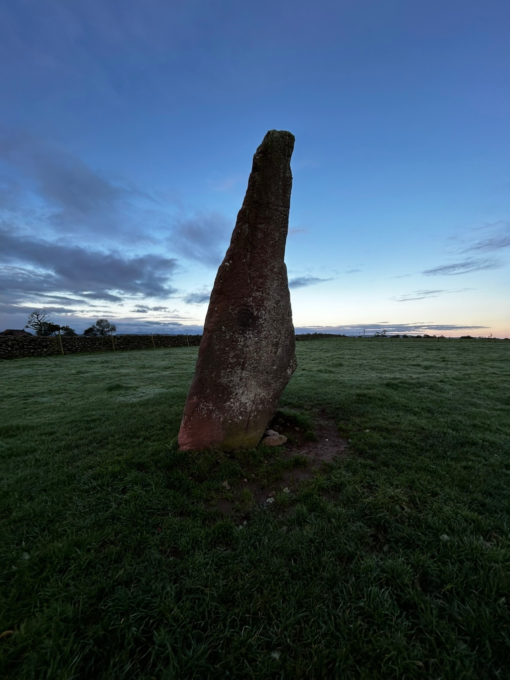 Tall, slender standing stone, possibly a menhir, in a grassy field. The stone is reddish-brown and appears weathered. The background is a clear, twilight sky with some clouds, and a low stone wall is visible in the distance.