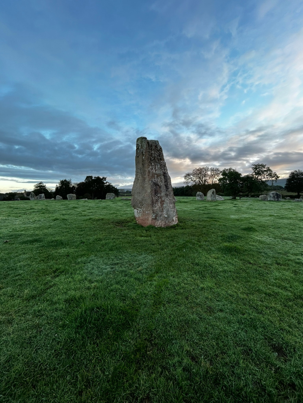 Single, tall, upright stone standing in a grassy field. Other, smaller stones are visible in the background, suggesting a stone circle or similar ancient monument. The sky is a clear, pale blue with streaks of cloud, and the grass appears slightly frosted.