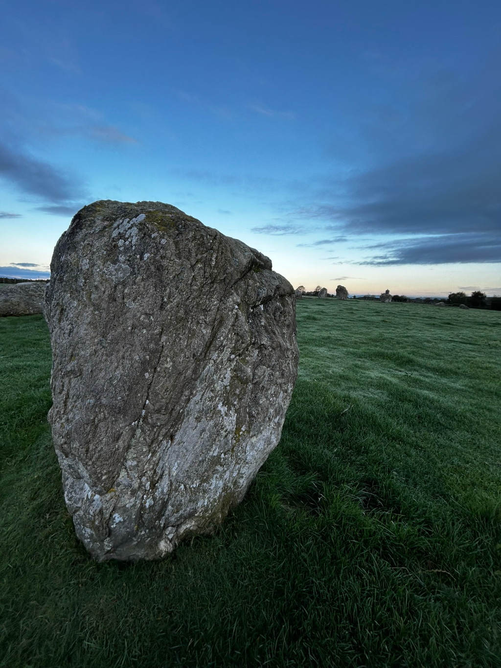 Large, grey boulder resting on a short, green, grassy field. Other, smaller boulders are visible in the distance, suggesting a stone circle or similar prehistoric monument. The sky is a clear, twilight blue, with some clouds visible. The overall impression is one of serenity and quietude, evoking a sense of history and the passage of time.