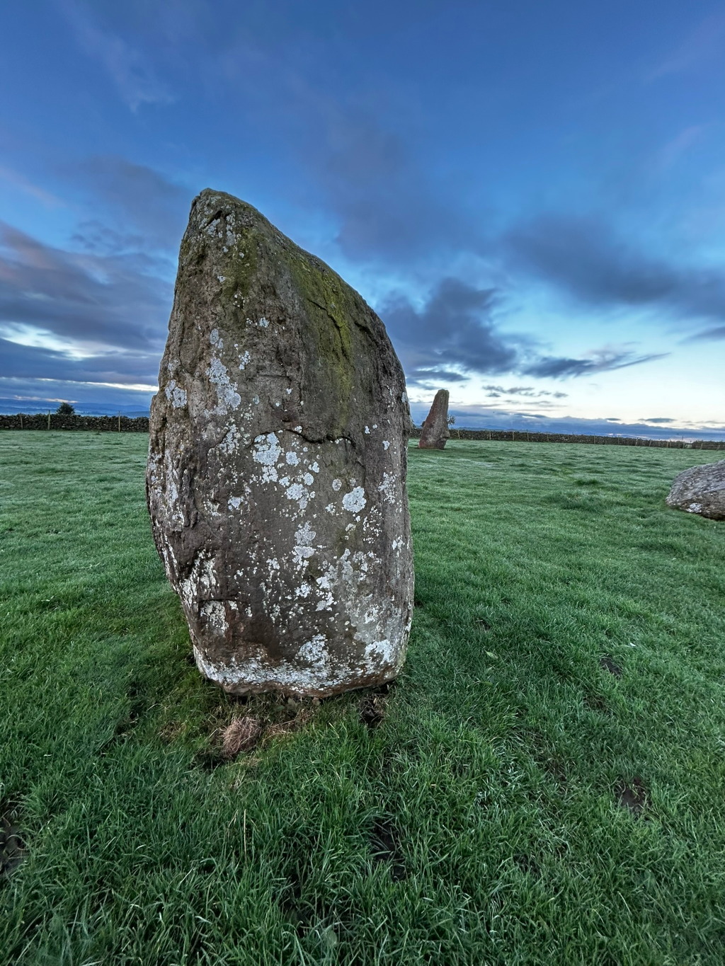 Large, moss-covered stone standing upright in a grassy field. Two smaller stones are visible in the background, suggesting a stone circle or similar ancient monument. The sky is a clear, twilight blue with some clouds.