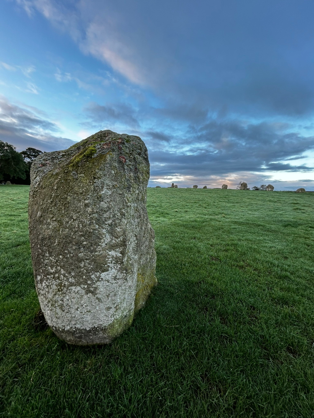 Large, moss-covered stone in the foreground, positioned in a grassy field. In the background, a line of smaller stones is visible, suggesting a stone circle or similar ancient monument. The sky is a mixture of clouds and blue, hinting dawn. The overall mood is serene and peaceful, with a focus on the contrast between the single prominent stone and the more distant, smaller stones.