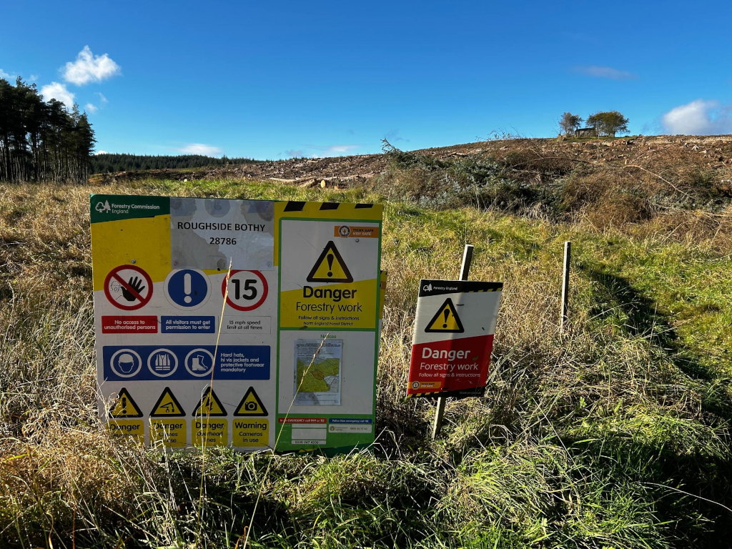 Landscape altered by forestry work, with a significant area of cleared land in the background. In the foreground, there are two prominent signs warning of ongoing forestry work and associated dangers. The larger sign, more detailed, lists several safety precautions, including speed limits, mandatory protective gear, and restrictions on unauthorised access. The smaller sign reiterates the danger of forestry work. The overall scene conveys a sense of both human intervention in a natural environment and the need for caution due to ongoing industrial activity.