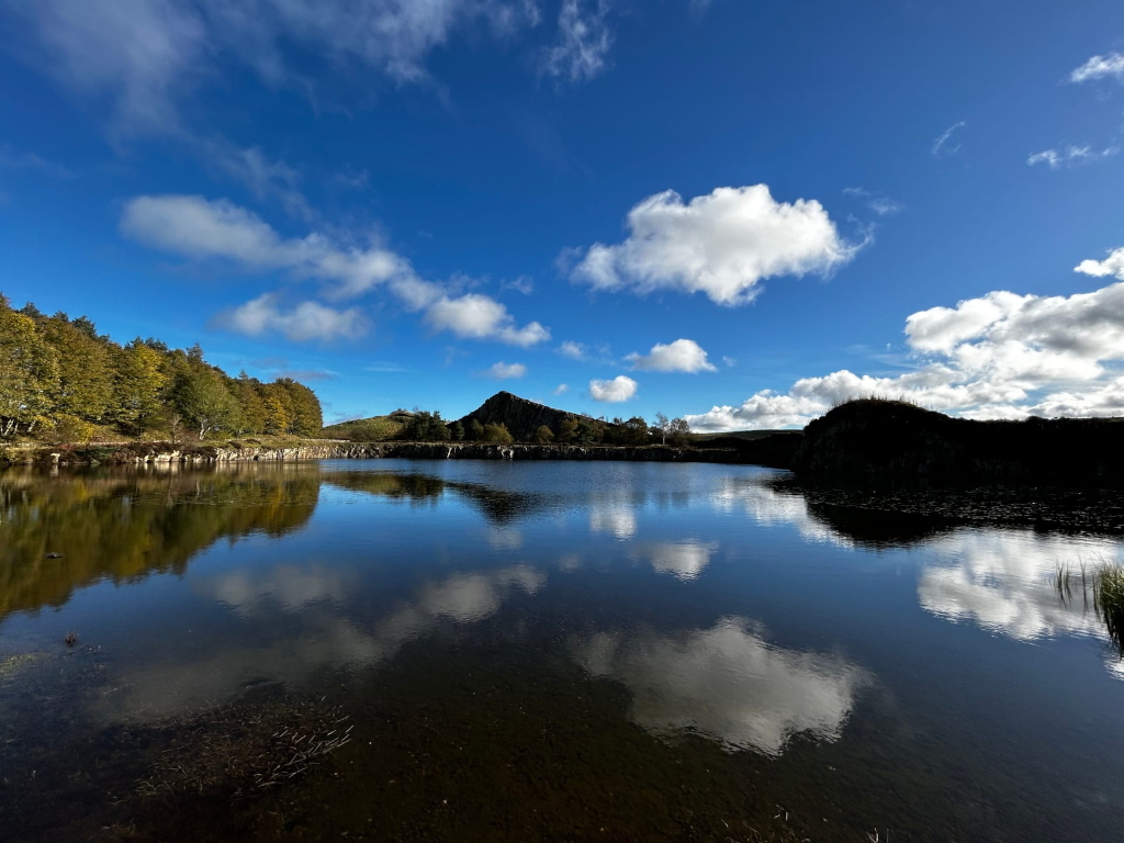 Serene lake reflecting a partly cloudy blue sky. The lake is surrounded by hills, one of which is a distinctive, dark-coloured rock formation. The far shore is lined with autumnal trees displaying varying shades of green and yellow.