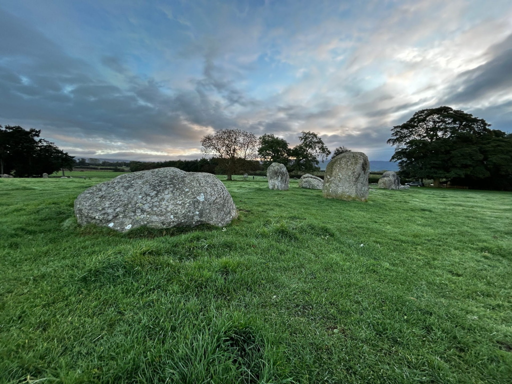 Serene landscape at dawn A short-grass field stretches across the foreground, leading to a small stone circle partially visible. A large, solitary boulder sits prominently in the foreground, while the remaining stones of the circle appear somewhat smaller and more weathered. Trees frame the background, with the sky a mixture of cloud and clear patches, suggesting a transitional time of day.