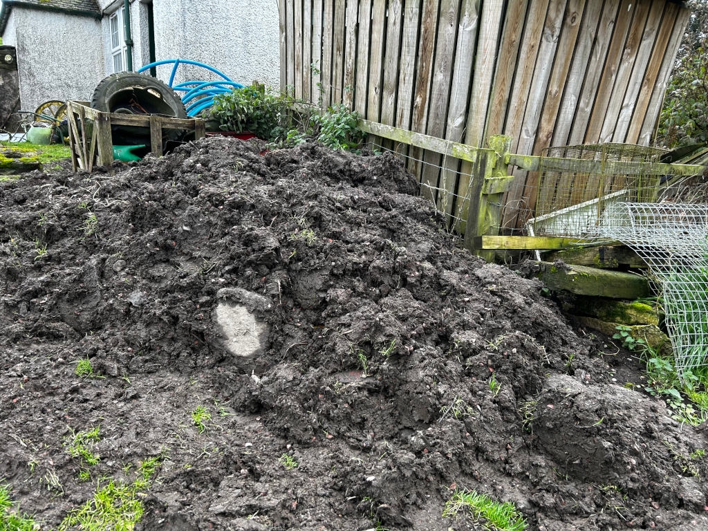 Large pile of dark, damp soil in a backyard setting. Behind the pile are parts of a wooden shed, fence, and some discarded items like a tire and what looks like a section of plastic pipe. The scene is somewhat cluttered and suggests recent gardening or landscaping activity. There are bits of grass and vegetation visible in the soil.