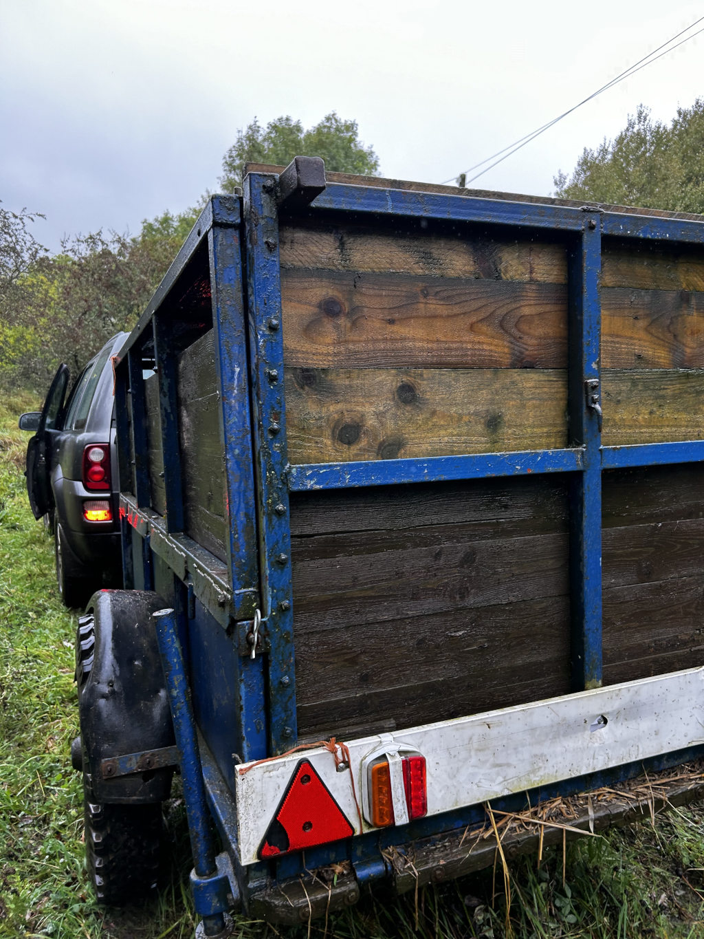 Rear view of a dark blue, weathered trailer hitched to a dark-coloured SUV. The trailer's walls are made of dark-stained wooden planks. The scene is set outdoors in a verdant, grassy area, with trees visible in the background, suggesting a rural or countryside location. The overall impression is one of rustic simplicity and perhaps a utilitarian purpose.