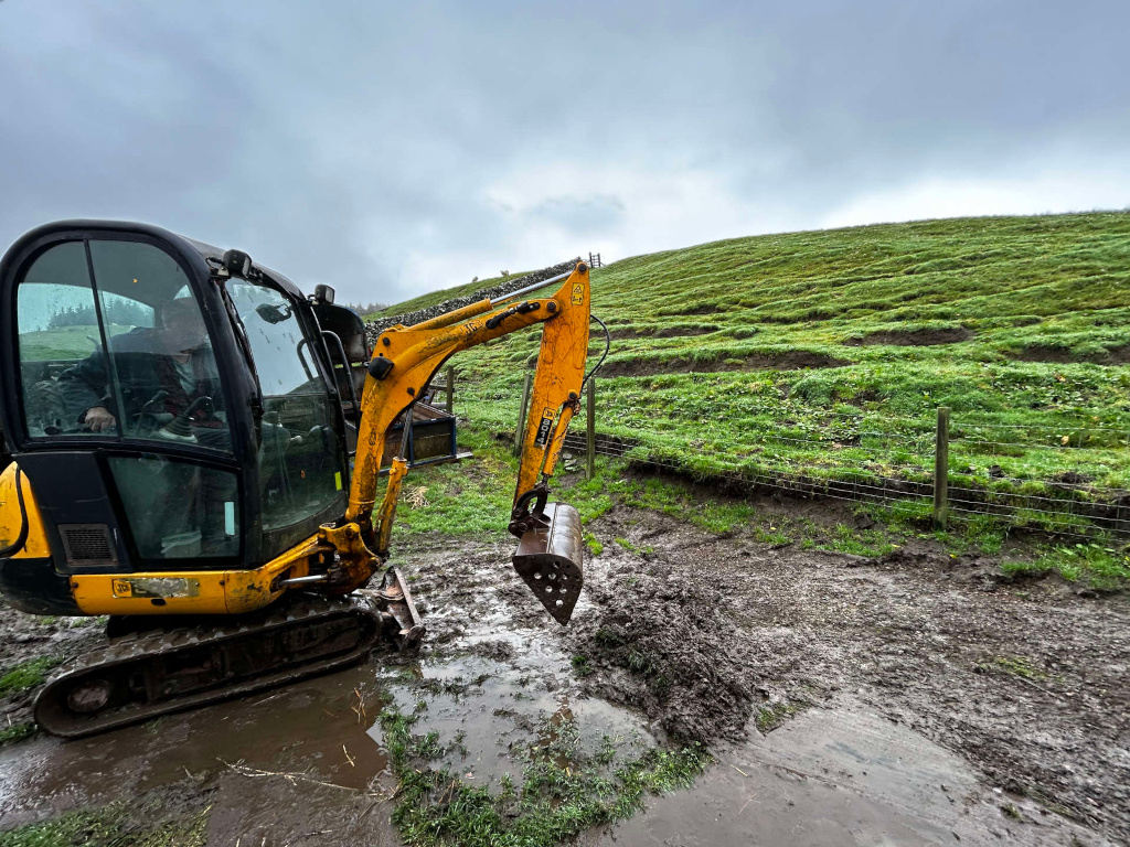 Yellow excavator, partially submerged in mud, operating near a grassy hillside with a wire fence. The excavator's driver, Charlie, is visible inside the cab. The overall setting appears to be a rural or agricultural area, possibly a farm or construction site, with overcast skies. The scene depicts the work of land shaping or related activity in challenging weather conditions.