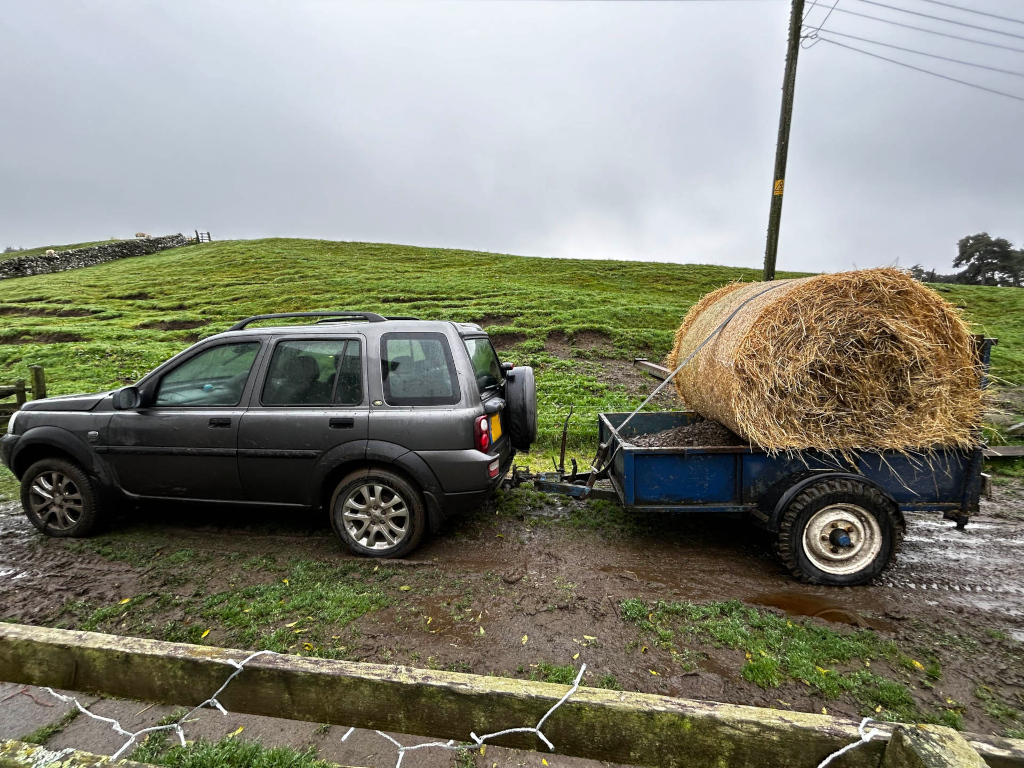 Dark grey Land Rover towing a small blue trailer carrying a large round bale of hay. The scene is set in a muddy farmyard or field, with a grassy hillside in the background under a cloudy sky. The overall impression is one of rural work or chores, possibly hay transportation on a farm.