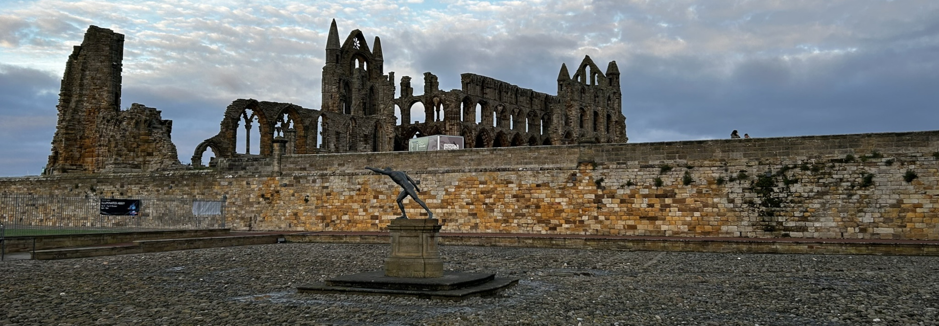 Whitby Abbey ruins in North Yorkshire, England. In the foreground is a stone pedestal holding a bronze statue of a discus thrower. The scene is set against a cloudy sky.