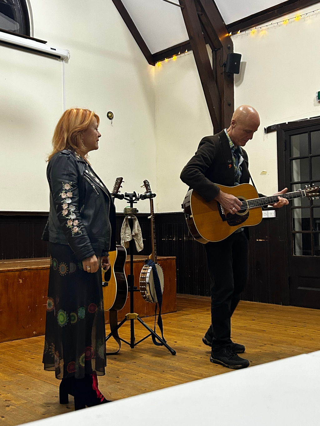 Man and a woman, possibly a musical duo, performing on a stage. The man is playing an acoustic guitar, while the woman stands beside him, possibly singing. Both are dressed in casual yet stylish attire. The setting appears to be a small, possibly community hall with wooden floors and exposed beams. Musical instruments, including a guitar, banjo, and microphone stand, are visible on the stage. The overall atmosphere is intimate and suggests a live acoustic performance.