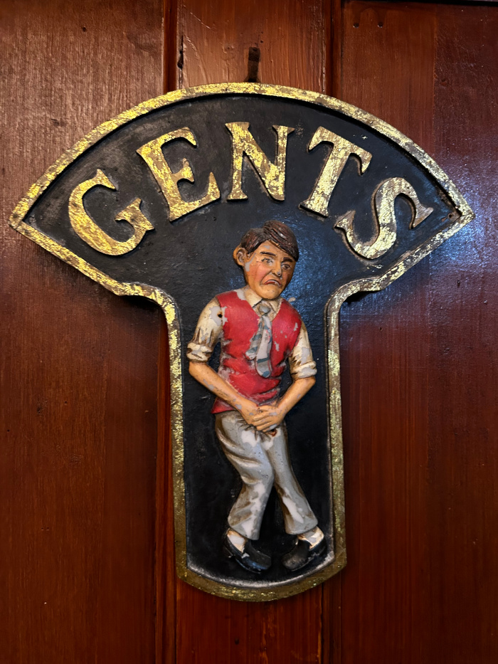Vintage gents' restroom sign. It's a dark-coloured, possibly cast iron or metal sign, shaped like a stylised T, with the word GENTS in gold lettering at the top. The main body of the sign features a three-dimensional relief sculpture of a man in a red sweater vest and light-colored trousers, appearing to be in a slightly uncomfortable, possibly urgent, posture related to needing to use the restroom. The overall style is humorous and slightly whimsical, typical of older pub or public house signage.