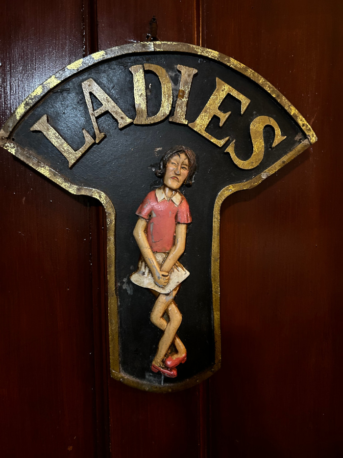 Vintage ladies' restroom sign. It's a dark-coloured, fan-shaped plaque with the word LADIES in gold lettering at the top.  Below the lettering is a three-dimensional carved figure of a woman in a dress, depicted in a crouching posture suggesting she needs to use the restroom. The overall style is whimsical and slightly humorous.