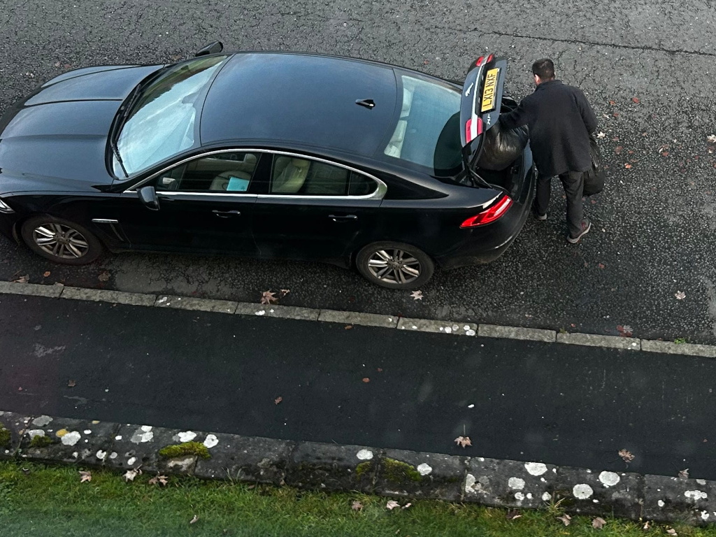 High-angle, long shot of a person placing a large black garbage bag into the trunk of a black Jaguar sedan parked on a residential street. The trunk is open, and the person is positioned behind the car. The street is bordered by a low stone curb and a strip of grass. The overall scene is mundane and unremarkable.