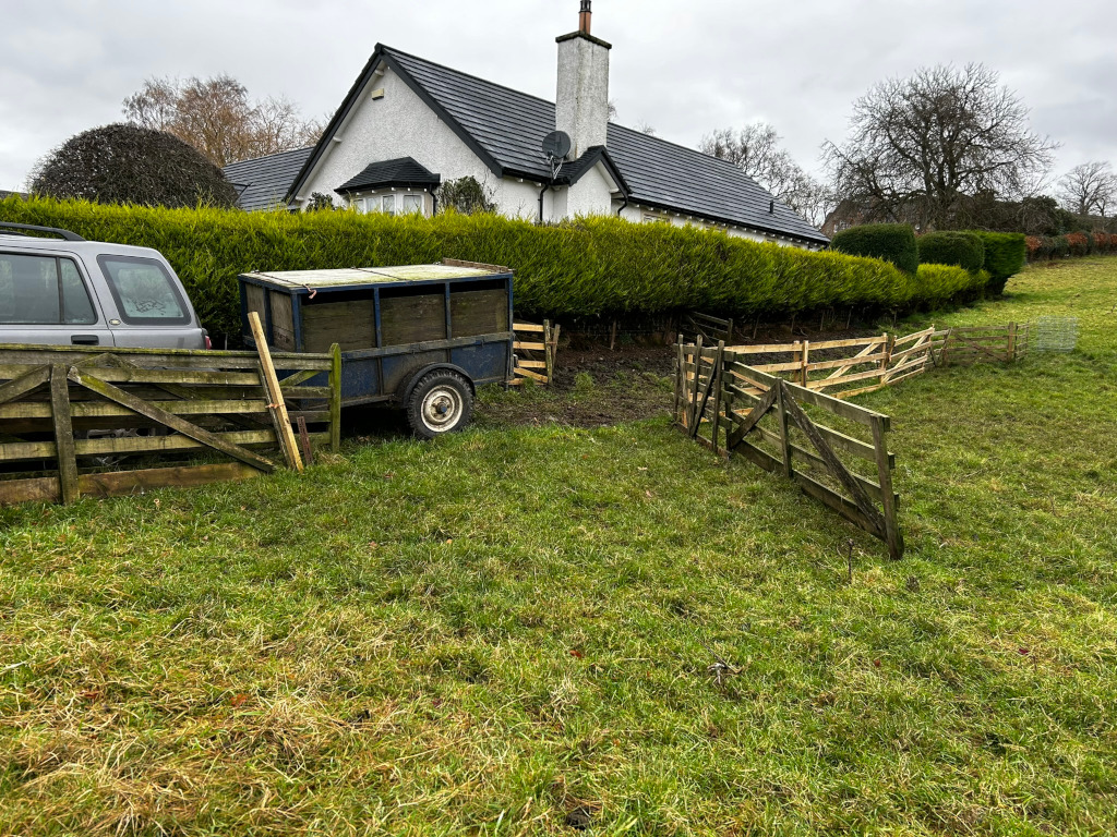 Rural scene with a small, white cottage nestled behind a neatly trimmed hedge.  In the foreground, there's a weathered, blue animal trailer parked next to a rustic wooden fence. A section of the fence forms a V-shape, possibly directing livestock. A silver SUV is partially visible behind the trailer.