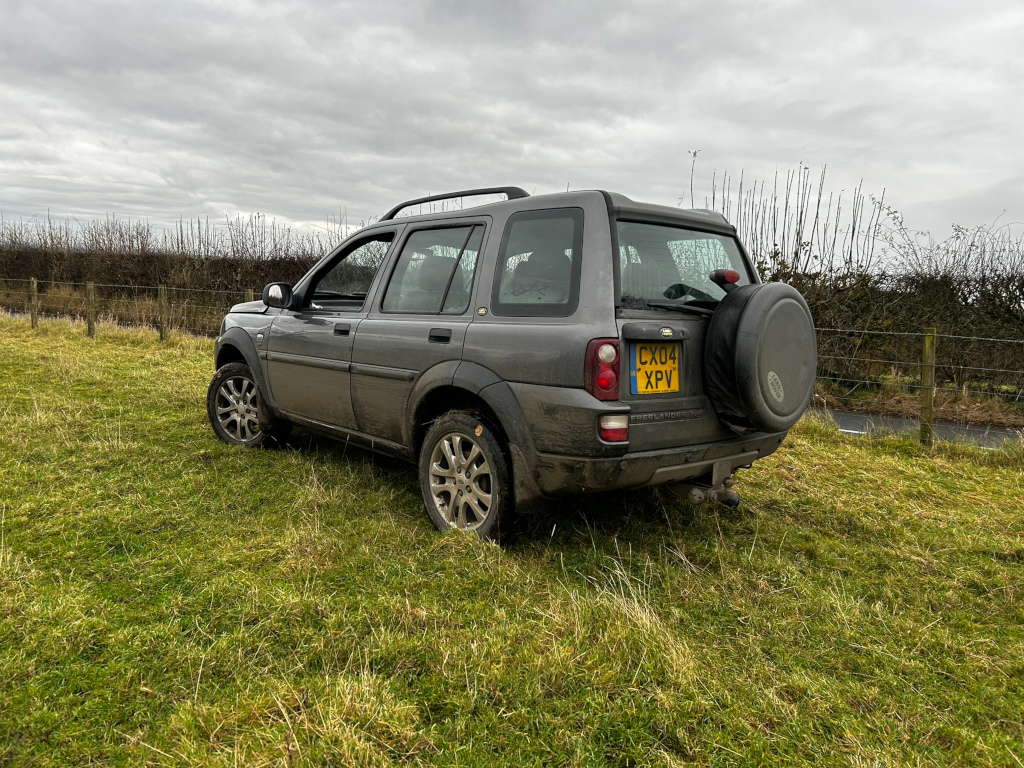 Grey Land Rover Freelander parked on a grassy field next to a fence. The sky is overcast. The vehicle is angled slightly away from the camera, showing its rear and side. A spare tire is visible mounted on the rear door.