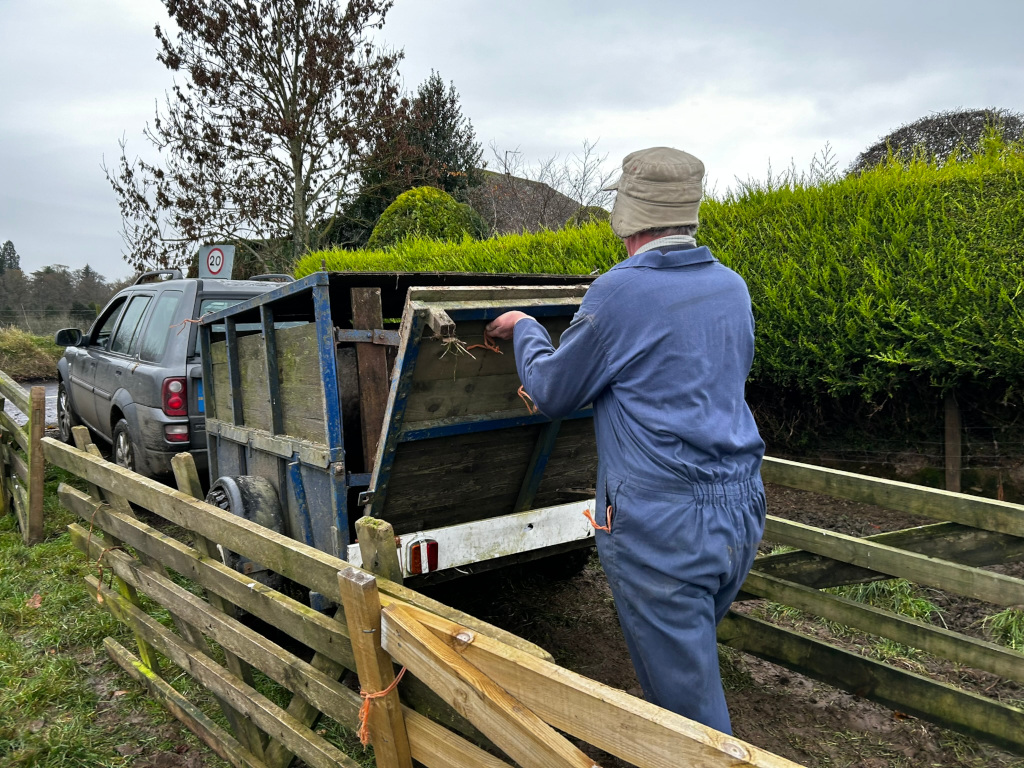 Charlie in blue overalls and a beige hat, seen from behind, loading what appears to be a trailer full of turf or sod.  The trailer is attached to a dark-grey Land Rover. The scene is set in a rural area, with a wooden fence and greenery in the background. The overall impression is of farm work or landscaping.