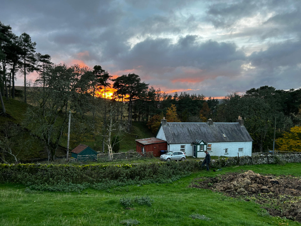 White cottage nestled in a rural landscape. A person is tending to a pile of manure in a garden in front of the house. A small car is parked near the cottage. The background features a hillside covered in trees, and a dramatic sunset with orange and gray clouds fills the sky. The overall scene is peaceful and evokes a feeling of rural tranquillity.