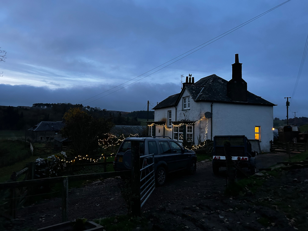 White two-story house in the countryside at twilight. String lights are strung along the exterior of the house. A dark-coloured SUV and a smaller utility vehicle are parked in the driveway. The scene is peaceful and evokes a feeling of rural tranquillity. A farm building is partially visible in the background. The overall mood is calm and slightly melancholic due to the dim light and the late hour.