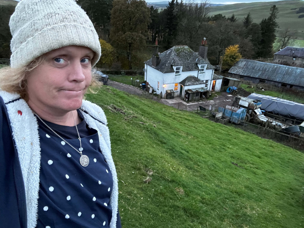 Leonie wearing a cream-coloured knit hat and a navy blue polka dot shirt under a cream-coloured fleece jacket, standing on a grassy hillside. Leonie is looking directly at the camera with a somewhat pensive or slightly unhappy expression. In the background is a small, white farmhouse and farm buildings, situated in a rural setting with rolling hills and trees. The overall mood is peaceful yet slightly melancholic.