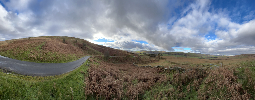 Panoramic view of a rural landscape, possibly in the UK. It features a winding asphalt road curving around a heather-covered hill. The foreground is dominated by dry, brown bracken and grasses, while a stone wall runs across the middle ground, separating the closer hill from a flatter expanse of land that stretches to the horizon. The sky is a mix of fluffy white clouds and patches of blue.