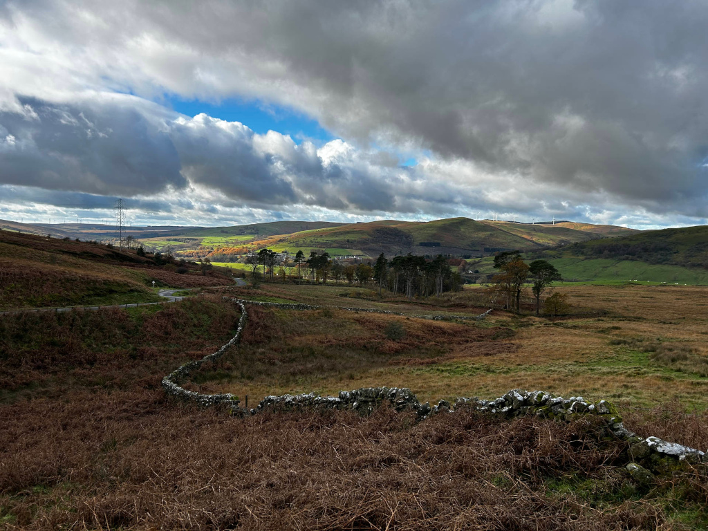 Pastoral landscape under a dramatic sky. The foreground features dry, brown grasses and a low stone wall winding through the terrain. A road curves gently in the mid-ground, leading towards a small cluster of trees and a village nestled in a valley. Rolling hills extend into the distance, with wind turbines visible on a ridge in the background. The sky is a mix of dark, heavy clouds and patches of blue, creating a moody yet beautiful atmosphere.