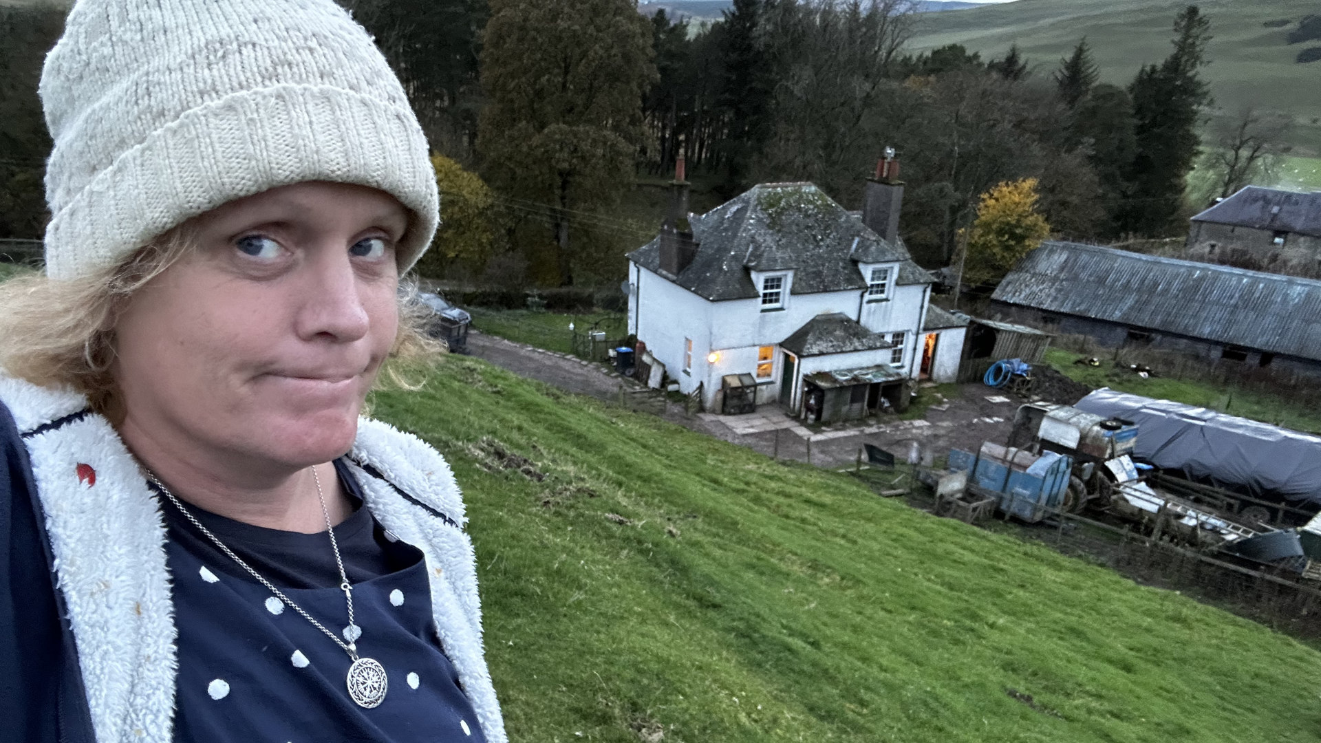 Leonie wearing a cream-coloured knit hat and a navy blue polka dot shirt under a cream-coloured fleece jacket, standing on a grassy hillside. In the background is a white farmhouse and several outbuildings, suggesting a rural setting. The overall mood appears to be somewhat pensive or contemplative.
