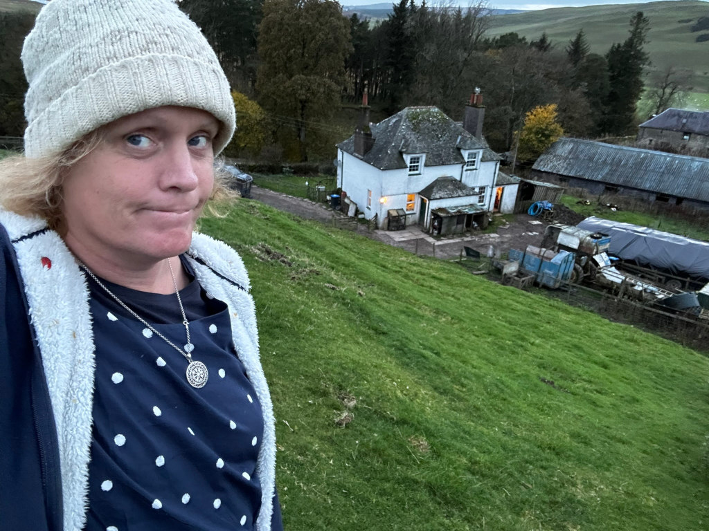 Leonie wearing a cream-coloured knit hat and a navy blue polka dot shirt under a cream-coloured fleece jacket, standing on a grassy hillside. In the background is a white farmhouse and several outbuildings, suggesting a rural setting. The overall mood appears to be somewhat pensive or contemplative.