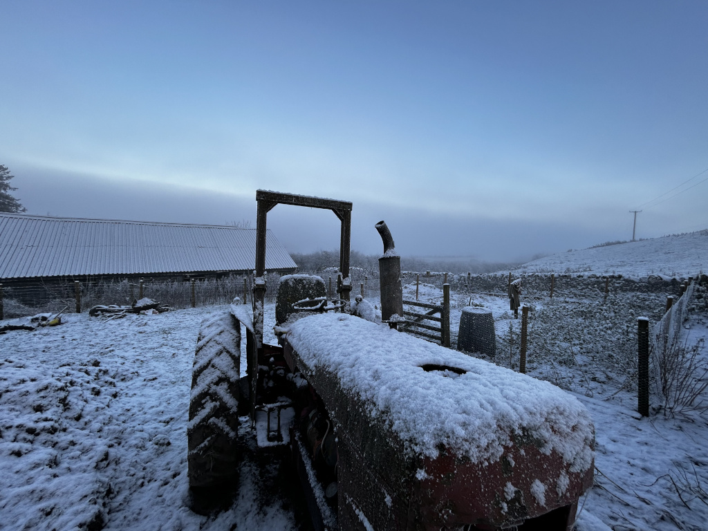 Snow-covered tractor parked in a farmyard. A snow-covered barn is visible in the background, along with a partly foggy landscape. The scene evokes a sense of quiet rural life in winter.