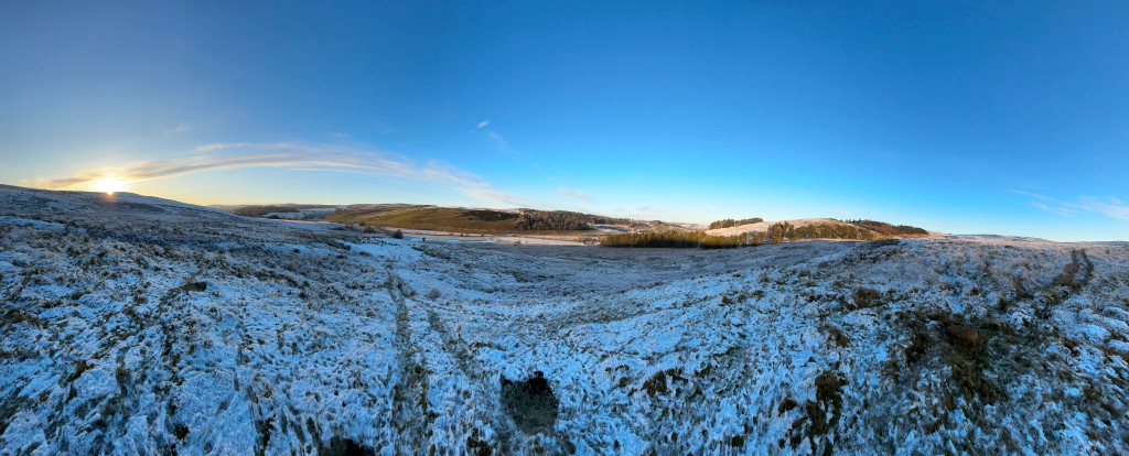 Panoramic view of a snow-covered landscape at sunrise. The foreground shows a field sparsely covered with low vegetation, dusted with a light layer of snow. Tire tracks are visible in the snow. In the distance, rolling hills extend to the horizon under a clear, vibrant blue sky. The sun is low on the horizon, adding a warm glow to the scene. The overall feeling is one of serene, wintry beauty.