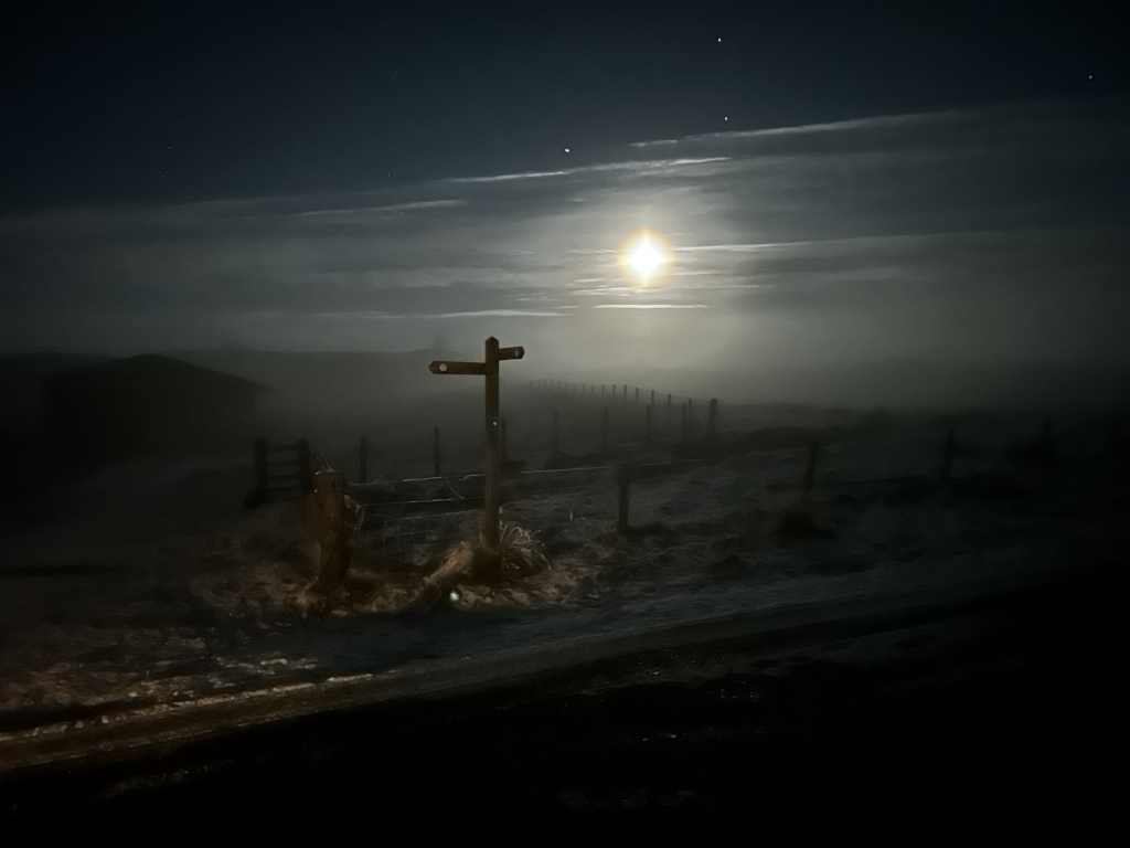 Solitary wooden signpost resembling a cross standing at a fence line in a misty, snowy landscape under a bright moonlit night. The atmosphere is mysterious and somewhat melancholic, emphasised by the low light, fog, and the isolated nature of the scene. The overall feeling is one of quiet solitude and possibly a sense of journey or pilgrimage.