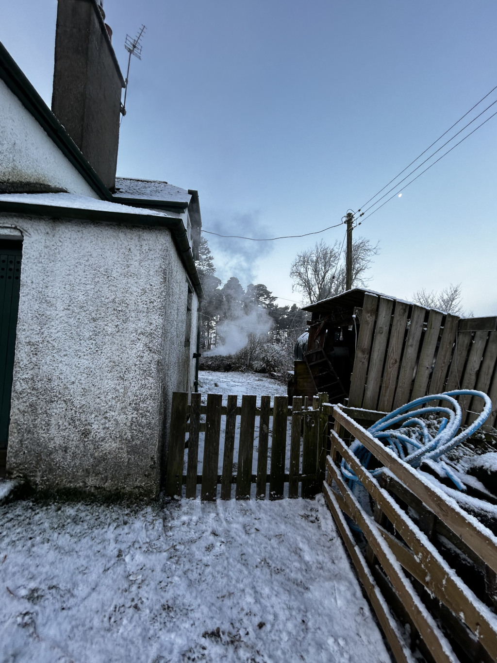 Snow-covered backyard scene. A whitewashed building, possibly a cottage, is in the foreground, with steam rising from its side, suggesting a heating system or perhaps a water source. A wooden fence and gate partially obstruct the view of a small yard, beyond which are trees and more structures, partially obscured by the snow and distance. The sky is clear and pale blue. 