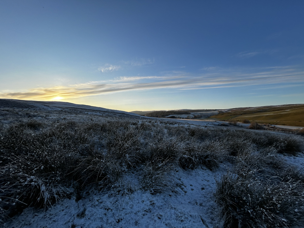 Tranquil winter landscape at either sunrise or sunset. The foreground shows a gently sloping hill covered in low-lying vegetation dusted with a thin layer of snow. The mid-ground and background reveal a broader valley, with rolling hills extending to the horizon. The sky is a clear, vibrant blue with subtle clouds, and the sun is low on the horizon, casting a warm glow.