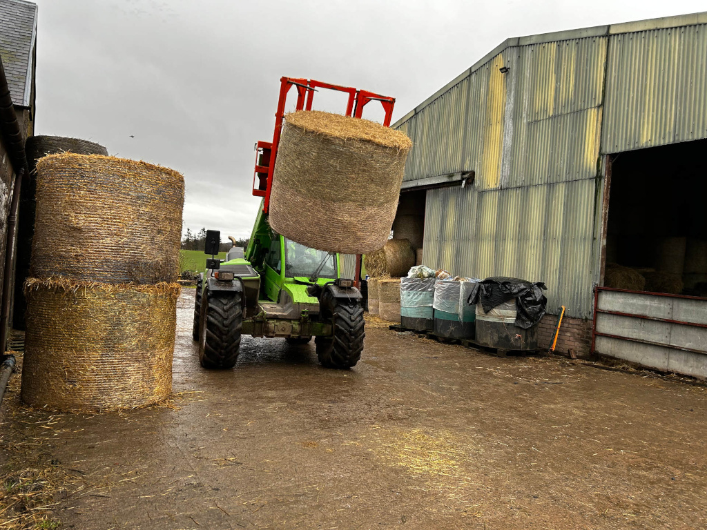 Green telehandler lifting a large round bale of hay in a farmyard. Two more round bales of hay are stacked beside a stone building. A corrugated metal barn is in the background along with several large black trash containers. The scene is overcast and damp. The overall impression is one of agricultural work and storage.