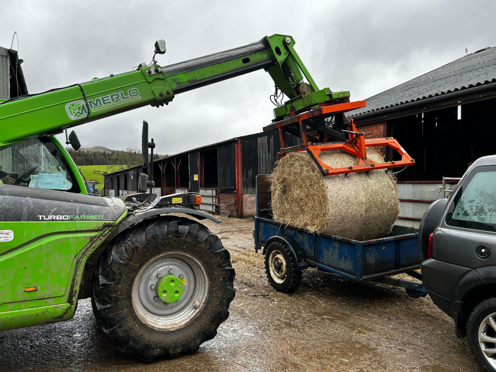 Green Merlo TurboFarmer telehandler lifting a large round bale of hay and placing it onto a small blue trailer towed by a dark-grey Land Rover. The scene takes place in a farmyard, with farm buildings and a hillside in the background under an overcast sky. The overall impression is one of routine farm work.